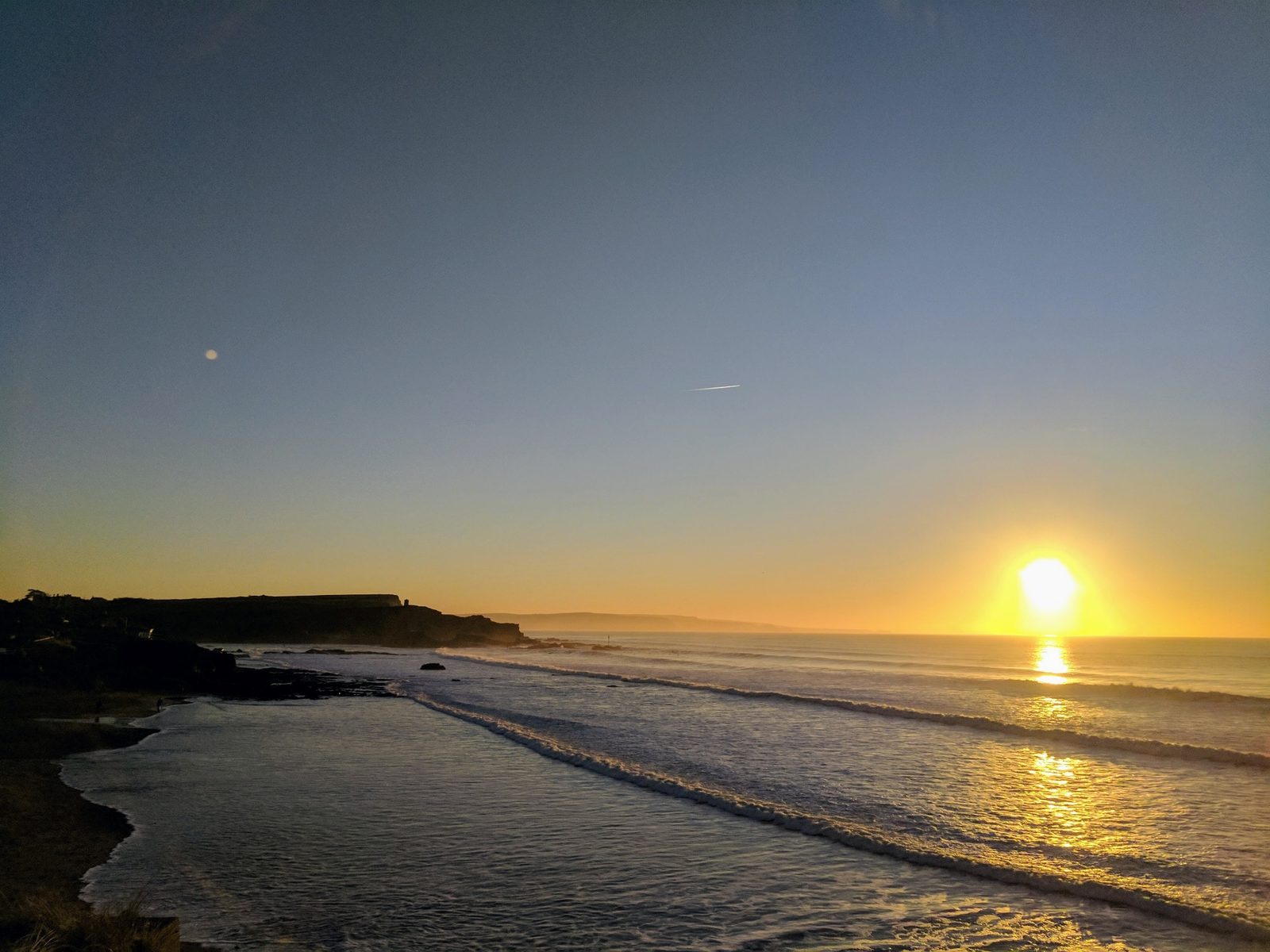 A crescent moon and setting sun over a Cornish coastline at dusk, waves rolling onto the beach