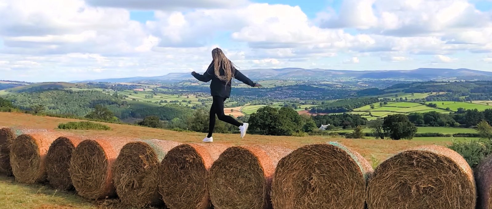 A person running along a line of round hay bales above a patchwork valley